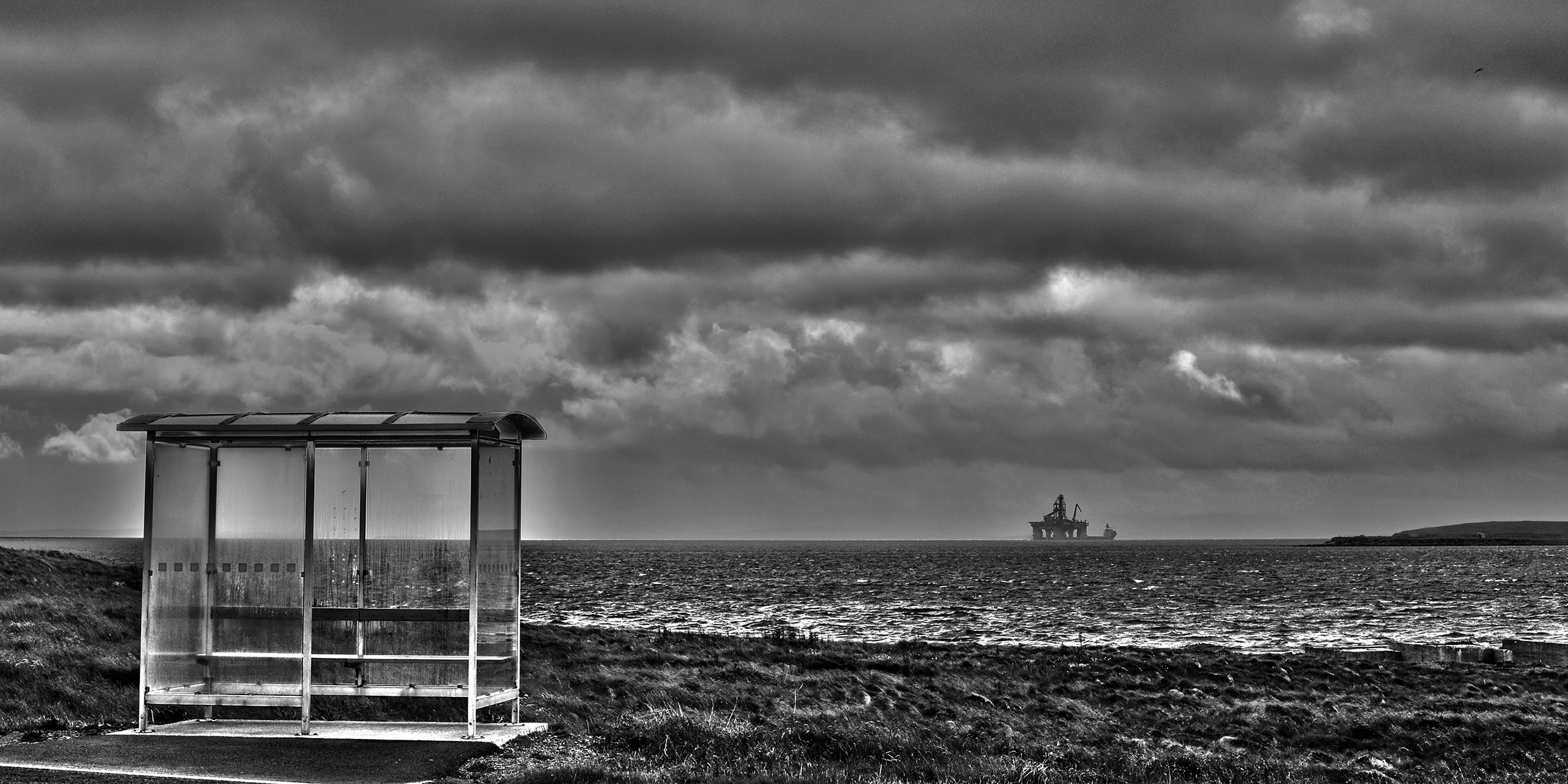 Et av bildene på utstillingen. Sort-hvitt foto av et busskur med hav og tunge skyer i bakgrunnen. Bakerst skimtes en oljeplattform.