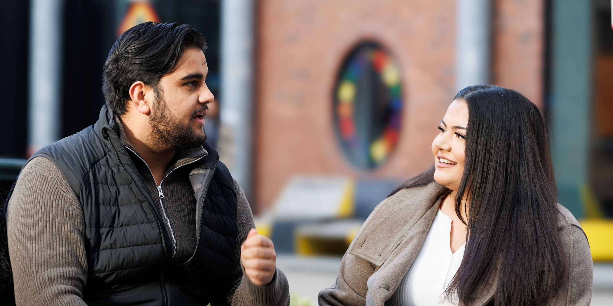 Two students, a boy and a girl, sit on a bench and talk to each other.