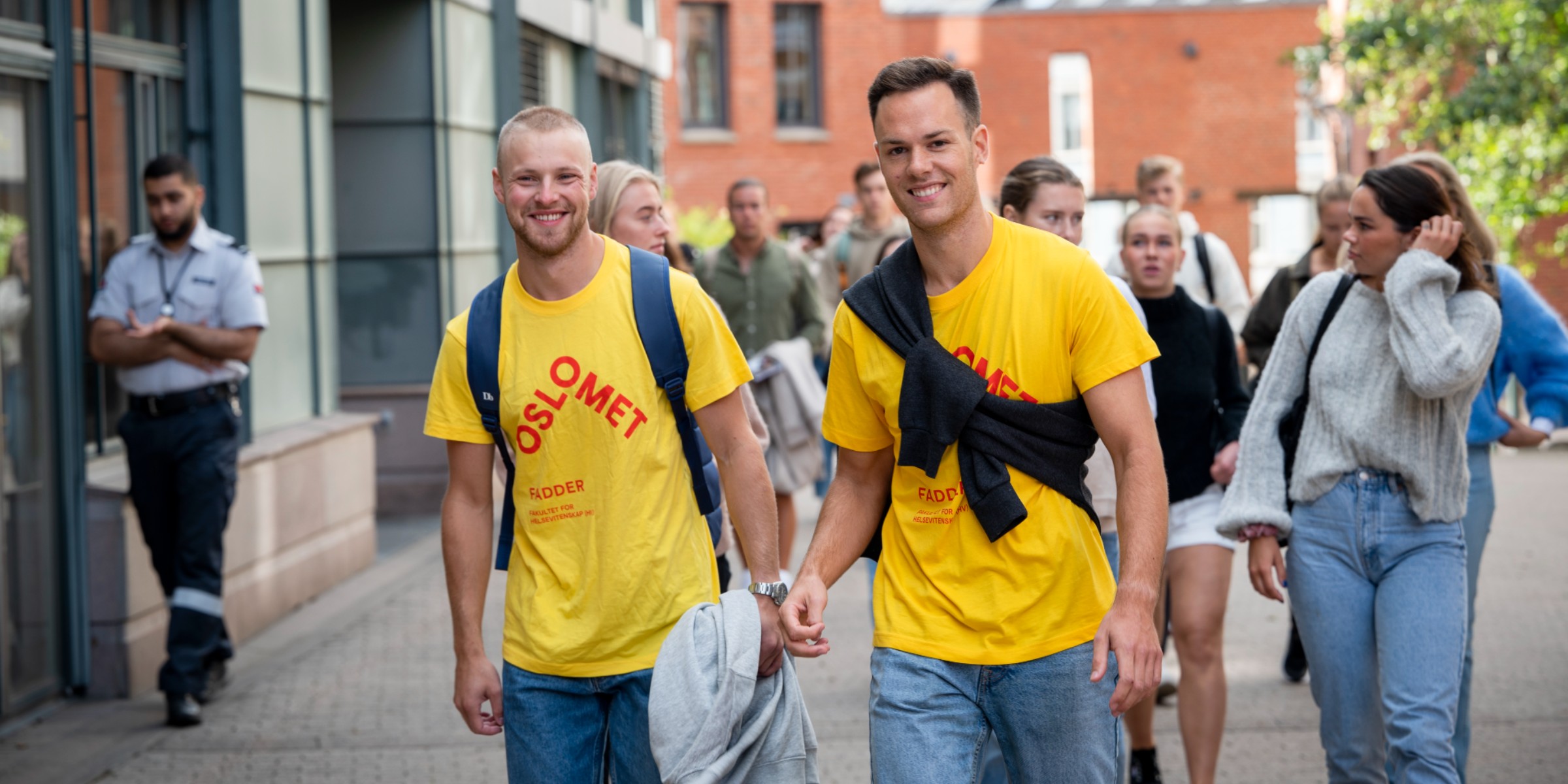 Two male students in buddy t-shirts smiling to camera with a group of new students walking behind them.